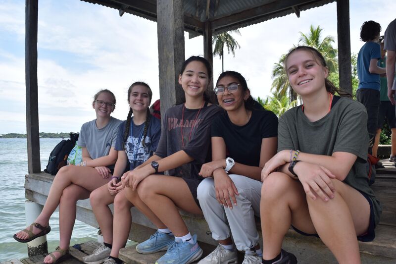 The image shows five young women sitting on a wooden dock. They are smiling and appear to be enjoying themselves. The background includes water, trees, and a cloudy sky. They seem to be on a trip or vacation together.