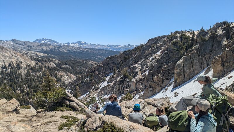 The image shows a group of people resting on a rocky outcrop, possibly during a hike. They are surrounded by a mountainous landscape with patches of snow. The sky is clear and blue, suggesting a sunny day. The terrain appears rugged and natural, with trees and exposed rock formations.