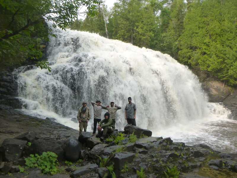 A group of five people stands on rocks in front of a large, cascading waterfall. The waterfall fills the background, surrounded by lush green trees. The people are positioned at the base of the falls, appearing small in comparison to the natural spectacle. The scene is captured on a cloudy day, with the light reflecting off the rushing water.