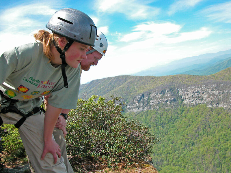 Two people wearing helmets stand on the edge of a cliff, looking down at the landscape below. The person in the foreground has blonde hair and is wearing a green t-shirt and beige pants. The other person is standing behind them. The background features a mountainous landscape with trees and a cloudy sky. They seem to be enjoying the view or preparing for an adventurous activity.