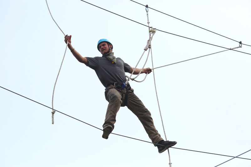 A man is carefully walking across a high ropes course. He is wearing a blue helmet and a grey t-shirt, with dark trousers and shoes. He is holding onto a rope with his left hand for balance. The ropes course is set against a pale sky, and the man appears focused on maintaining his balance as he navigates the challenge.