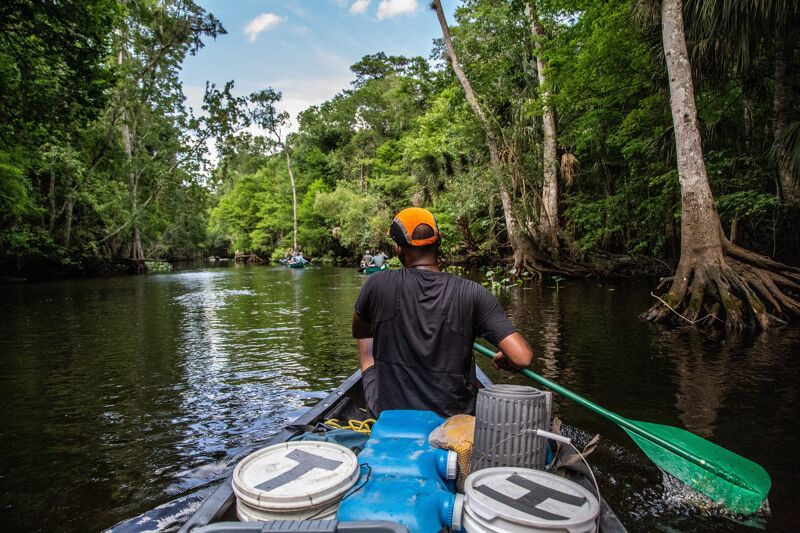 A person wearing a black shirt and an orange cap is paddling a canoe through a river surrounded by lush green trees. The river reflects the sky and the surrounding vegetation. The canoe has some white buckets with black letters on them. The scene is peaceful and serene, capturing the beauty of nature.