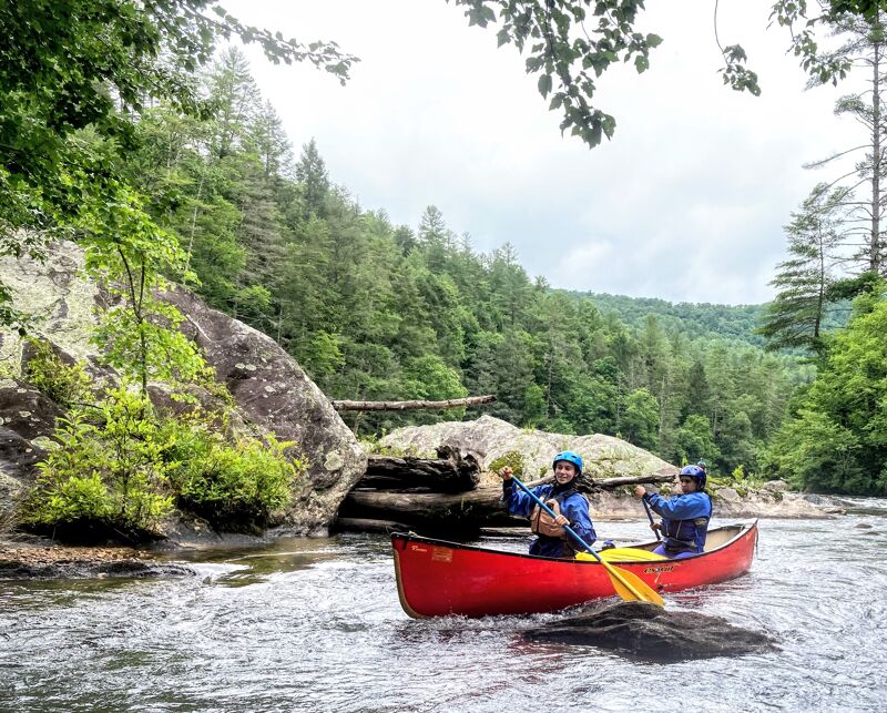 Two people are in a red canoe on a river. The person in the front is wearing a blue jacket and a blue hat and is holding a paddle. The person in the back is wearing a blue jacket and a helmet. The river is surrounded by trees and rocks. The sky is cloudy.