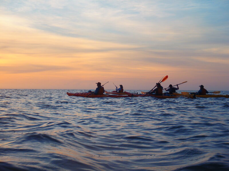 The image shows a group of people kayaking on the ocean during a sunset. The sky is a mix of orange, yellow, and blue, reflecting on the water's surface. The kayakers are silhouetted against the bright sky, and they appear to be paddling in the same direction. The water is choppy, with small waves visible in the foreground.