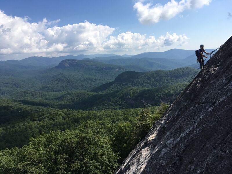 A person is rock climbing on a steep rock face. They are using ropes and gear for safety. In the background, there is a lush green forest covering rolling hills and mountains. The sky is blue with scattered white clouds, suggesting a clear day. The overall scene captures an adventurous outdoor activity in a scenic natural environment.