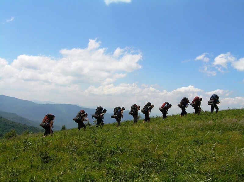 A group of hikers with large backpacks are walking along a grassy ridge under a blue sky with scattered clouds. The hikers are silhouetted against the bright sky, and the landscape includes rolling hills and mountains in the distance. The scene conveys a sense of adventure and the challenges of hiking in a natural environment.