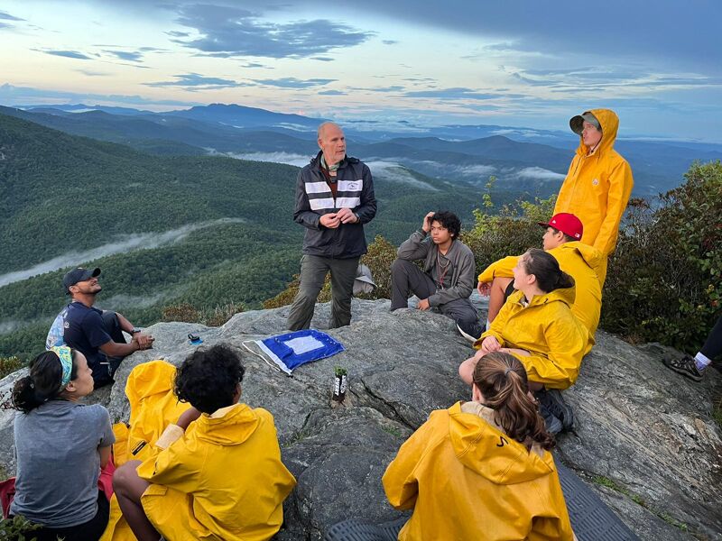 A group of people are gathered on a rocky mountaintop, some wearing yellow raincoats. A man in a dark jacket appears to be addressing the group, with mountains and a cloudy sky in the background. The setting suggests an outdoor educational or team-building activity.