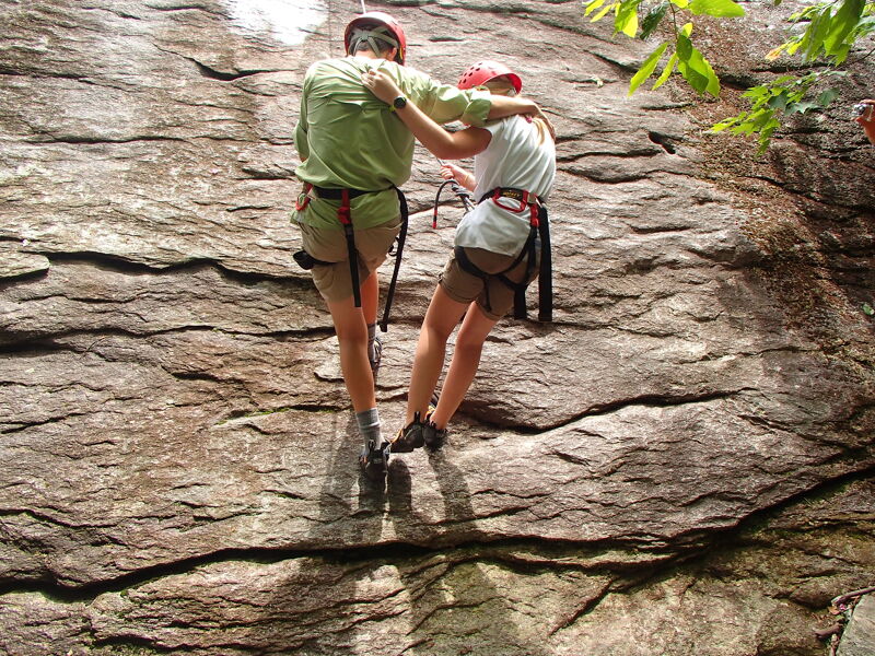 Two people are rappelling down a rock face. The person in the front is wearing a green shirt and red helmet, while the person behind them is wearing a white shirt and red helmet. Both are wearing harnesses and are attached to ropes. The rock face is textured and has some vegetation growing on it.