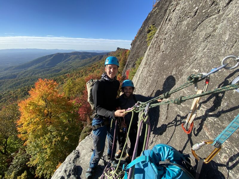 Two rock climbers are on a cliff face, geared up with helmets and harnesses. They are surrounded by climbing ropes and equipment. Behind them, a scenic view unfolds with mountains, trees displaying autumn colors, and a clear blue sky. The climbers appear to be taking a break or preparing for their next ascent.