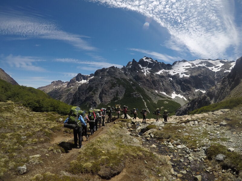 A group of hikers is trekking along a narrow path in a mountainous region. The hikers are carrying large backpacks, suggesting a multi-day trip. The landscape features rugged terrain with patches of vegetation and rocky outcrops. In the background, snow-capped mountains rise against a blue sky with scattered clouds, adding to the scenic beauty of the location.