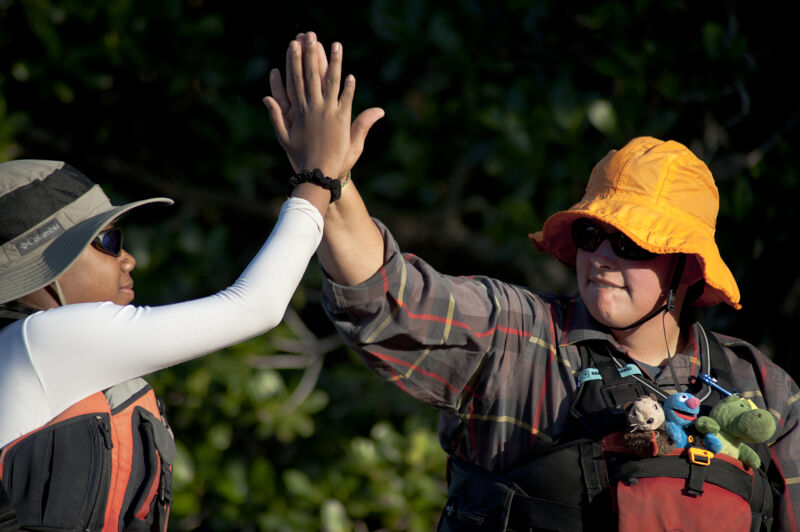 Two people wearing hats and life vests are giving each other a high five. The person on the left is wearing a grey hat and a white long-sleeved shirt under their vest. The person on the right is wearing an orange hat and a plaid shirt. They also have what appears to be a stuffed animal attached to their vest. The background is blurry, but it looks like they are outdoors with trees in the distance.