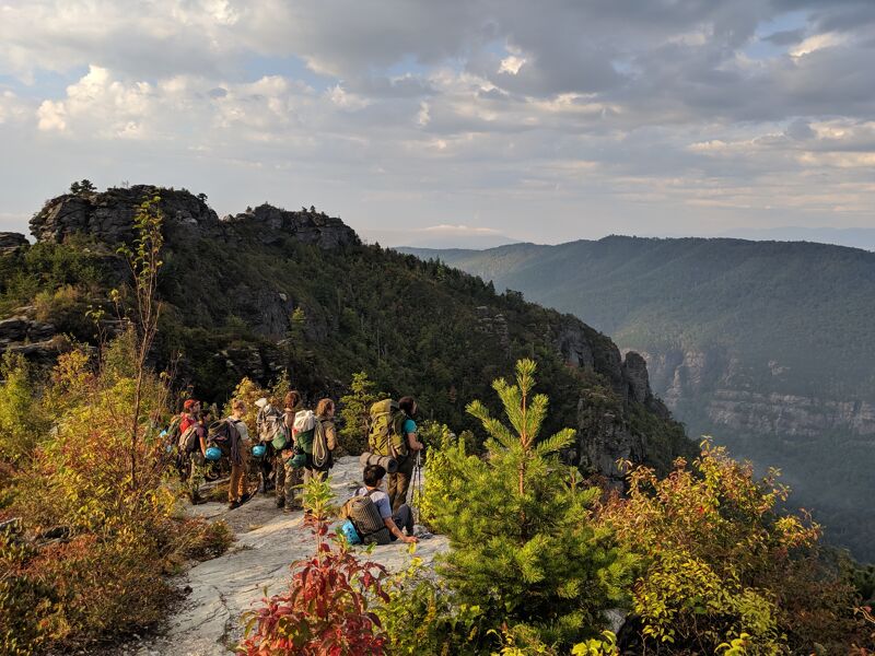 A group of hikers is resting on a rocky outcrop, likely a mountain summit or high point, enjoying a scenic view. They are surrounded by lush greenery, including trees and shrubs, with a backdrop of rolling hills and mountains under a partly cloudy sky. The landscape suggests a wilderness or natural park setting.