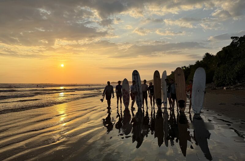A group of people are standing on a beach at sunset, holding surfboards. The sun is setting over the ocean, casting a warm glow on the scene. The people are silhouetted against the light, and their reflections can be seen in the wet sand. The surfboards are of various shapes and sizes, and some of the people are looking out at the waves.