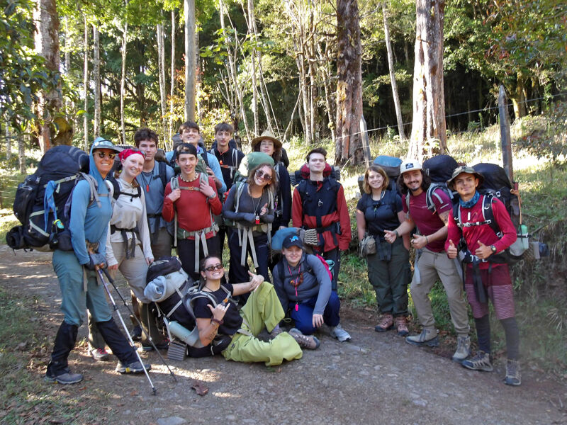 A group of about 15 people are standing on a dirt path in a wooded area. They appear to be hikers, as they are wearing backpacks and hiking gear. Some are smiling and looking at the camera. The background is a dense forest with tall trees. The lighting suggests it is daytime.