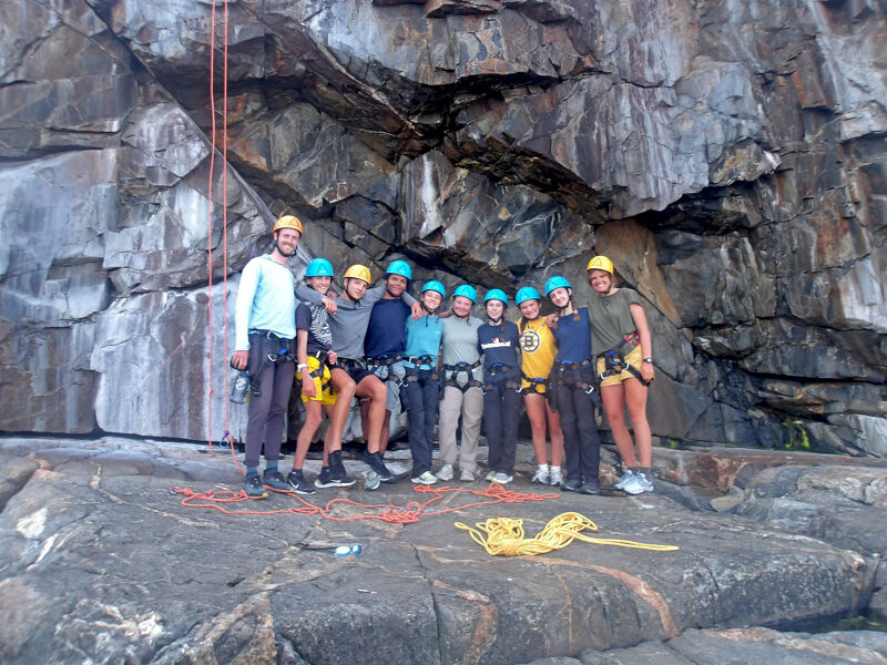 A group of about a dozen people are standing on a rocky surface in front of a large rock wall. They appear to be climbers, as some are wearing helmets and harnesses, and ropes are visible. The rock wall is gray and textured, with some dark patches. The people are dressed in a variety of casual clothing, and the overall scene suggests an outdoor adventure activity.