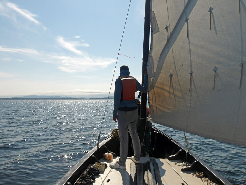A person stands on the bow of a sailboat, facing away from the viewer. They are wearing a life vest, light-colored pants, and a dark shirt. The sail is partially visible on the right side of the frame. The sea is calm with a distant shoreline visible under a blue sky with scattered clouds. The boat appears to be moving through the water.
