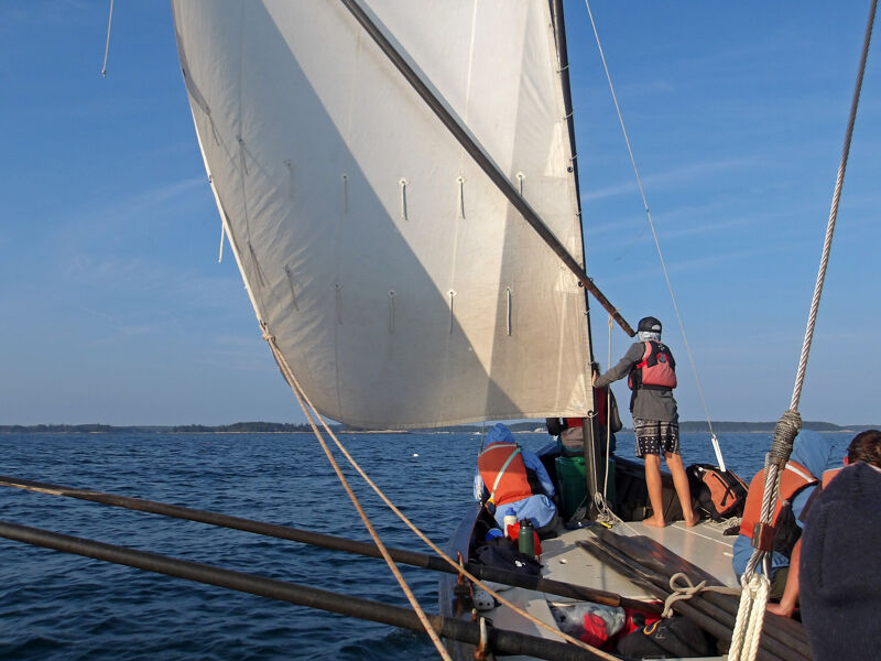 The image shows a sailboat on the water with several people on board. The boat has a large white sail that is full of wind. The people on the boat appear to be enjoying a day of sailing. The sky is blue and the water is calm. The overall scene is peaceful and serene.