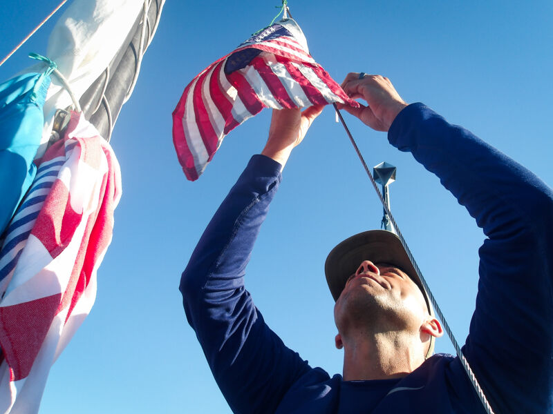A man is hoisting an American flag on a sailboat. He is wearing a dark blue long-sleeved shirt and a hat. The sky is clear and blue. There are other flags hanging from the mast of the sailboat. The man is looking up at the flag as he raises it.