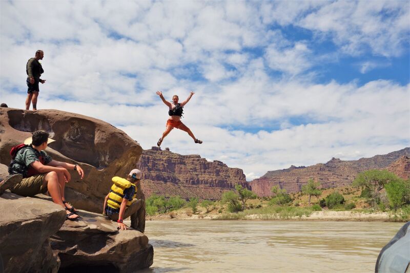 A group of people are enjoying a sunny day by a river. One person is captured mid-air, leaping from a rocky outcrop with arms outstretched. Another person stands atop the rocks, while two others are seated, observing the jumper. The river flows calmly, and the background features a scenic landscape of mountains and trees under a partly cloudy sky.