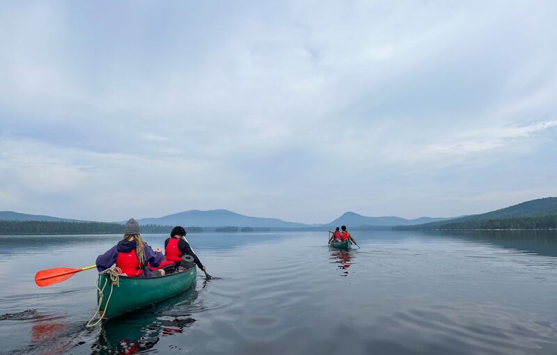 The image shows three people in canoes on a calm lake, surrounded by mountains and a cloudy sky. The person in the foreground canoe is wearing a red life jacket and paddling. The water is still, reflecting the sky and the distant shoreline. The overall scene conveys a sense of tranquility and outdoor adventure.