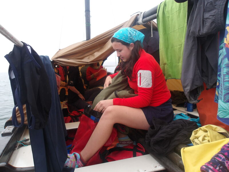 The image shows a young woman on a boat. She is wearing a red jacket, blue shorts, and a blue bandana. She is surrounded by clothes and other items. The boat appears to be covered with a makeshift shelter.