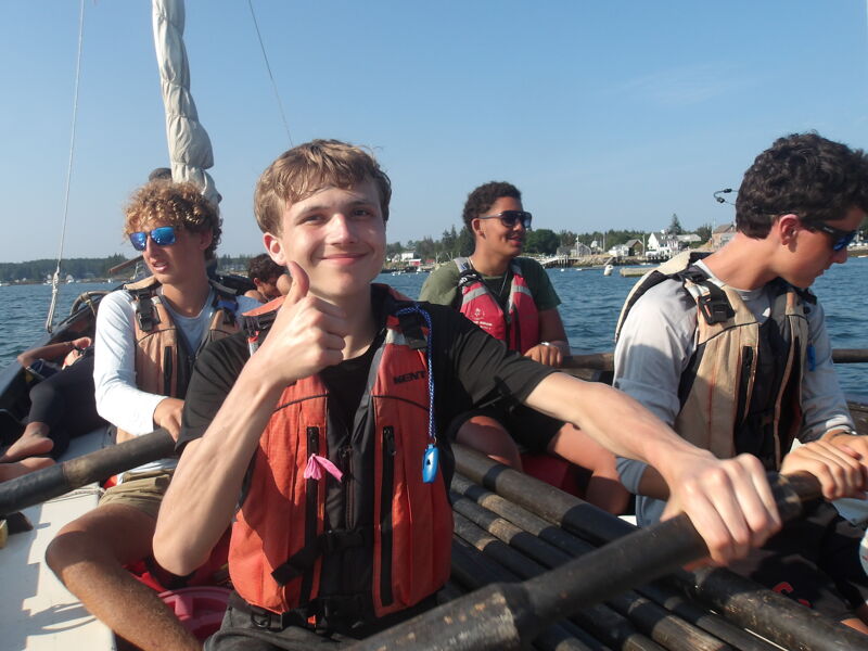 The image shows a group of young people on a boat. The person in the foreground is giving a thumbs up. They are all wearing life jackets. The background shows a body of water and some land. It looks like a sunny day.
