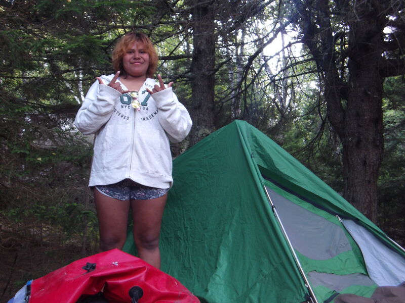 In the image, a person stands in front of a green tent in a forest. The person is wearing a white jacket and shorts, and they are making a hand gesture. There is also a red object in the foreground. The scene suggests a camping or outdoor activity.