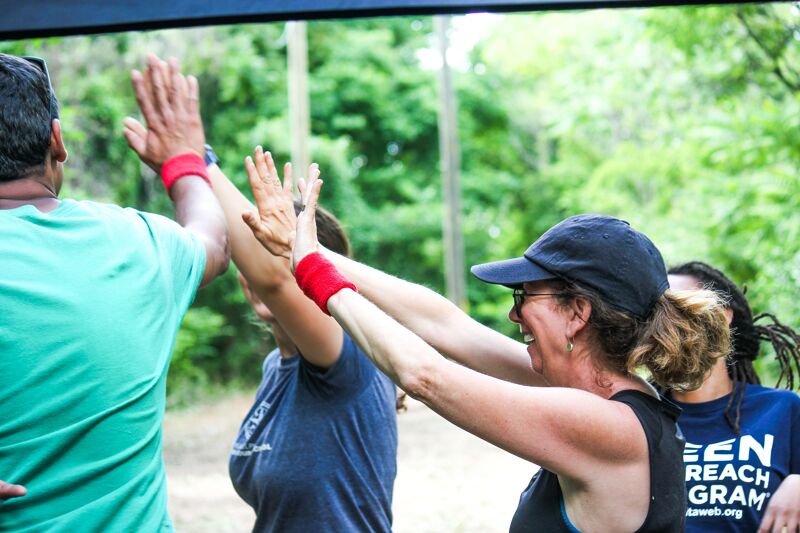 A group of people are giving each other high fives outside. The person on the left is wearing a green shirt. The person in the middle is wearing a blue shirt and a red wristband. The person on the right is wearing a blue shirt with white text. They are all smiling and appear to be in a good mood. The background is a blur of green trees.