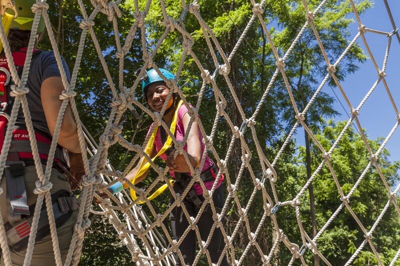The image shows two individuals participating in a ropes course or similar outdoor activity. The person in the foreground, wearing a helmet and safety harness, is navigating a rope net obstacle. The focus is on this person, who is smiling and appears to be enjoying the challenge. Another person, also equipped with safety gear, is partially visible to the left, seemingly assisting or participating alongside. The background features lush green trees, suggesting the course is set in a wooded area.