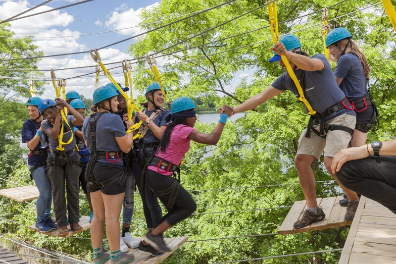 A group of people are participating in a ropes course. They are wearing helmets and harnesses and are attached to safety lines. The course is set up in a wooded area, and the participants are walking across a rope bridge. One person is reaching out to help another person across the bridge. The weather appears to be sunny.