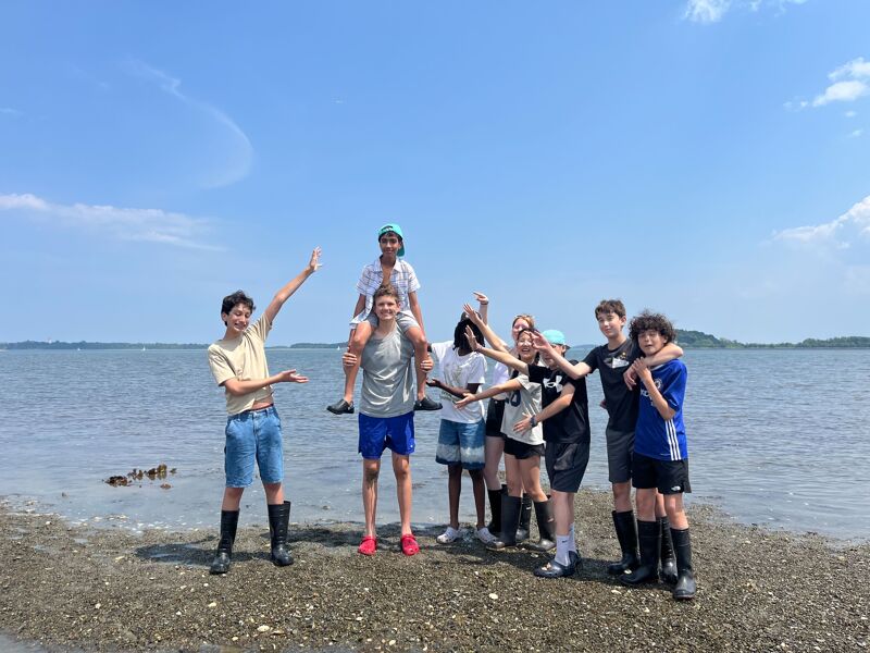 The image shows a group of young people posing on a beach on a sunny day. Some are standing, while others are sitting on shoulders. They are all smiling and appear to be enjoying themselves. The background features a calm sea and a clear blue sky.