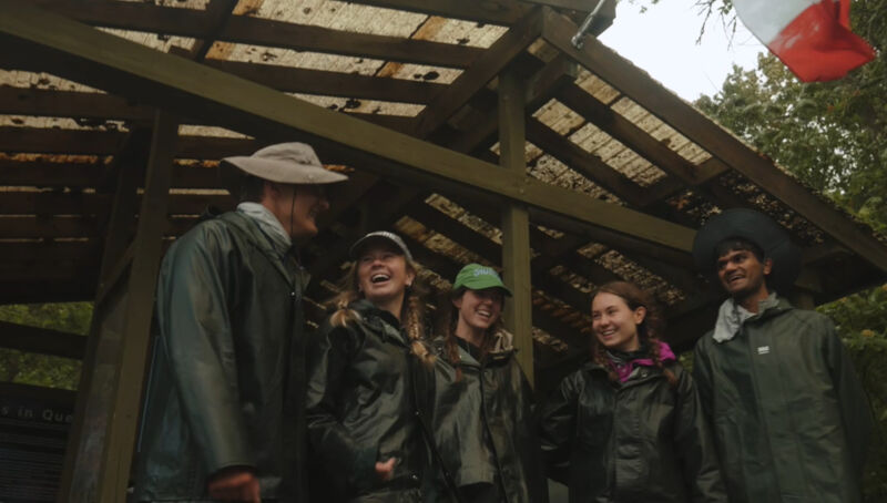 The image shows a group of five people standing under a wooden structure, possibly a shelter or shed. They are all wearing raincoats, suggesting it might be raining or the weather is wet. The people appear to be smiling and in good spirits. A flag, possibly Polish, is visible in the background, adding a cultural element to the scene.