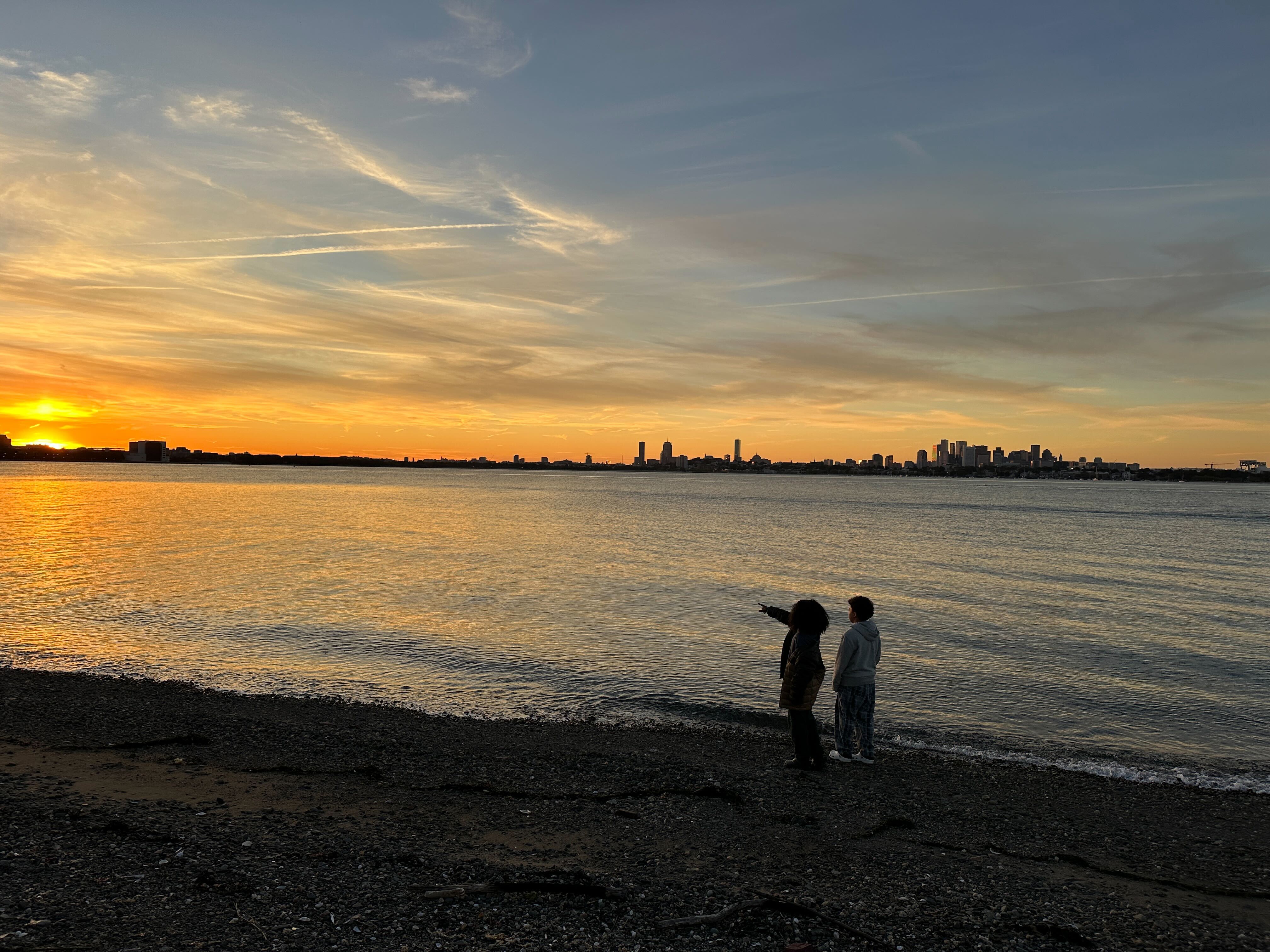 The image captures a serene sunset over a body of water, likely a sea or a large lake. Two figures stand on the shore, gazing towards the horizon where the sun is setting, painting the sky with vibrant hues of orange and yellow. A city skyline is visible in the distance, silhouetted against the colorful sky.