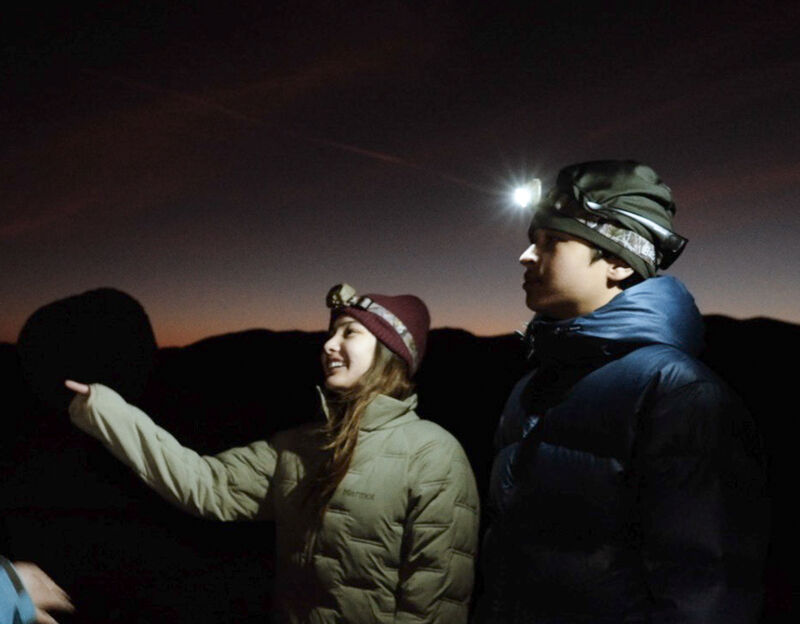 The image shows two people standing outside at dusk or dawn. Both are wearing winter hats and puffy jackets. The person on the right has a headlamp on, illuminating their face. The person on the left is pointing towards something in the distance. The background is dark, suggesting it is either early morning or late evening.