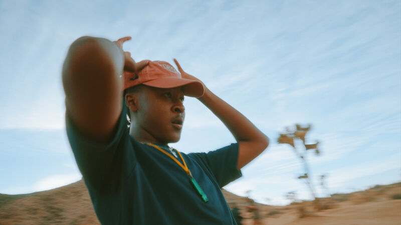 A man in a dark blue t-shirt and an orange cap stands in a desert landscape under a partly cloudy sky. He is holding his hands up near his head. In the background, there's a structure resembling a windmill or weather monitoring device. The overall scene suggests a sunny, possibly windy day in a remote, arid location.