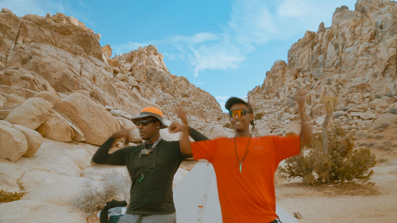 Two men are standing in a rocky, desert-like landscape with mountains in the background. The man on the left is wearing a black long-sleeved shirt and an orange hat, while the man on the right is wearing an orange t-shirt. Both men have their arms raised in the air, appearing to be celebrating or posing for a photo. The sky is blue with some clouds.