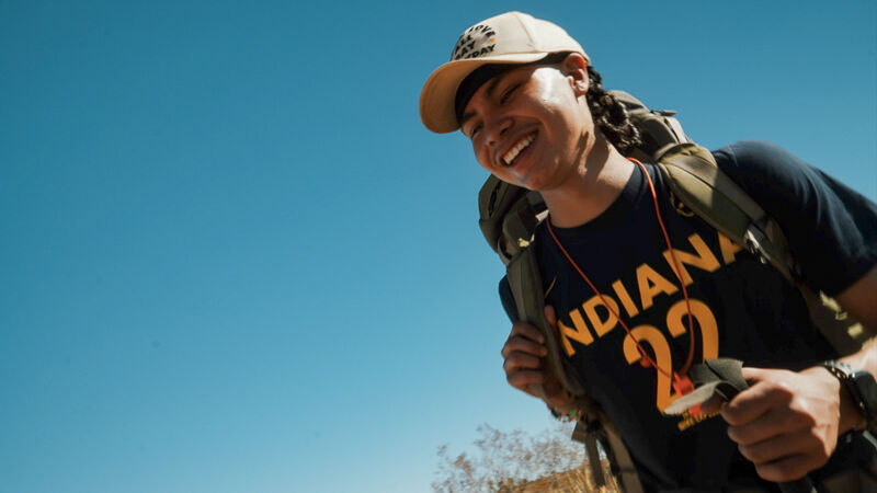 A person wearing a baseball cap and a backpack is smiling against a clear blue sky. They are wearing a dark blue t-shirt with "INDIANA 22" printed in gold. The person is holding something in their hands, possibly a compass or a small device. The overall impression is one of adventure and happiness in an outdoor setting.