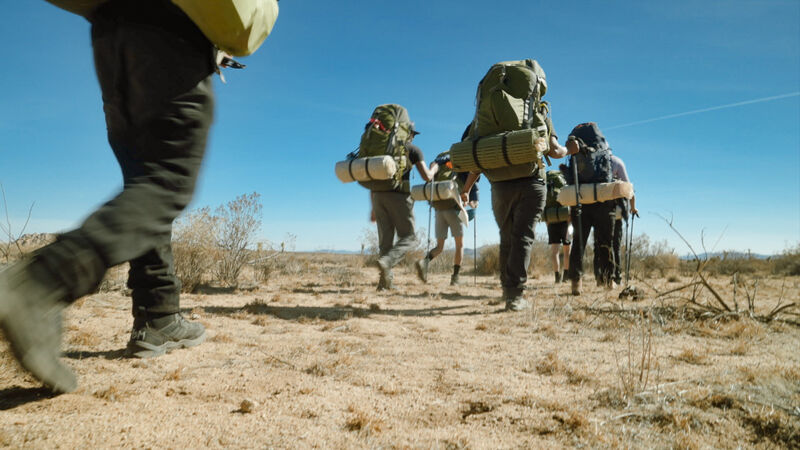 A group of hikers is trekking through a desert landscape under a clear blue sky. They are equipped with large backpacks and hiking poles, suggesting a multi-day or long-distance journey. The terrain is arid and sparsely vegetated, with dry grasses and shrubs dotting the sandy ground. The hikers are spread out, indicating a steady pace across the open expanse.