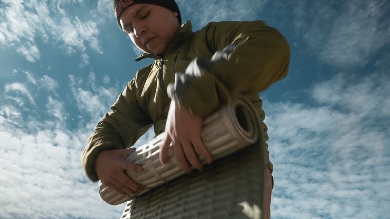 A person wearing a black beanie and a green jacket is holding a rolled-up sleeping pad against a backdrop of a blue sky with scattered clouds. The individual appears to be securing or adjusting the pad, with both hands firmly grasping it. The shot is taken from a low angle, emphasizing the person and the gear against the sky.