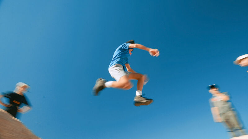 A man is captured mid-air, seemingly jumping from one surface to another. The background is a clear, bright blue sky. Other people are visible in the scene, with one appearing to be in motion blur, suggesting movement. The overall impression is one of action and athleticism.