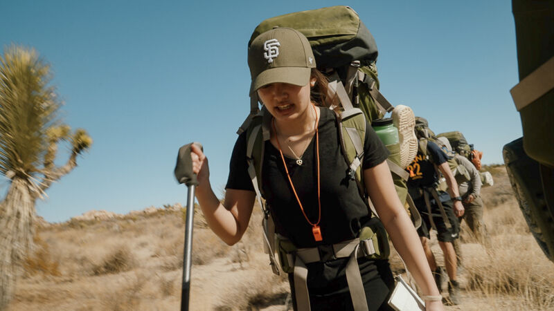 A woman wearing a baseball cap and a large backpack is hiking in a desert environment. She is holding a hiking pole and appears to be part of a group, as other hikers are visible behind her. The landscape is arid with sparse vegetation under a clear sky.