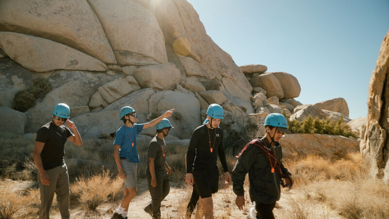 A group of five people are hiking in a rocky, desert-like environment. They are wearing helmets, suggesting they might be rock climbing or engaging in some other outdoor adventure activity. The landscape features large boulders and sparse vegetation under a clear, sunny sky. The group appears to be following a trail, with one person pointing ahead, possibly indicating the direction or a point of interest.