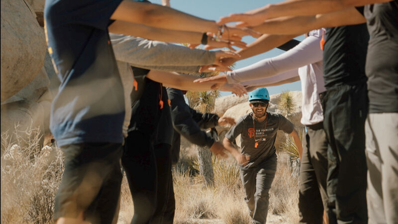 A group of people are standing in a circle with their arms outstretched, while a man in a blue helmet runs towards them. The group appears to be in a desert or rocky area, with sparse vegetation visible in the background. The man running towards the group is smiling and appears to be excited to join them.