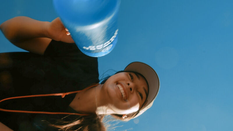 The image shows a woman from a low angle, holding a blue water bottle against a clear blue sky. She is smiling and wearing a black shirt and a baseball cap. The composition emphasizes the vastness of the sky and the woman's upward gaze, creating a sense of optimism and connection with nature. The bright lighting and vibrant colors contribute to a cheerful and refreshing atmosphere.