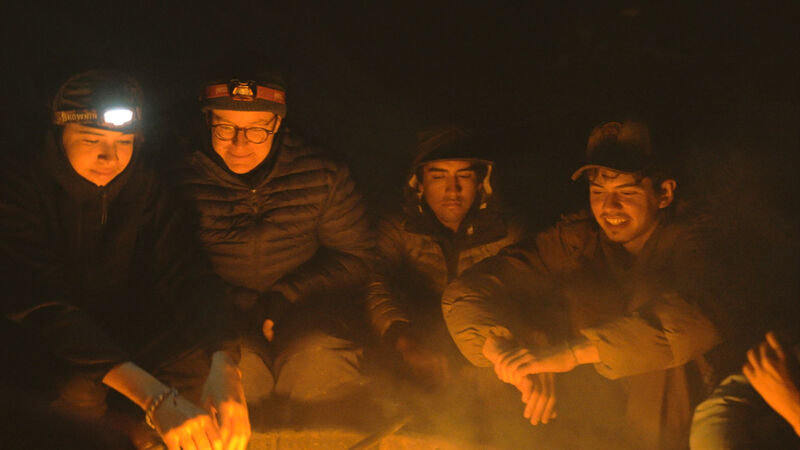 The image shows four people sitting around a campfire at night. They are all wearing headlamps or hats, and they appear to be warming their hands by the fire. The fire is casting a warm glow on their faces, and the overall scene is one of camaraderie and warmth in the darkness. The setting seems to be outdoors, possibly camping or a similar activity.
