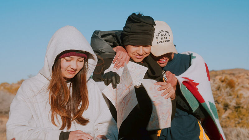 Three young people are huddled together outdoors, looking at a map. One person, wearing a white hooded jacket, is looking down at something in their hands. Another person in the middle is wearing a black beanie and holding the map. The third person, wearing a baseball cap and wrapped in a blanket, has their arms around the other two. They appear to be friends or family on an adventure.