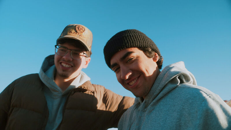 The image shows two young men smiling at the camera. The man on the left is wearing glasses, a brown jacket, and a baseball cap. The man on the right is wearing a black beanie and a grey hoodie. The background is a clear blue sky. They appear to be outdoors, possibly enjoying a sunny day. Both men have a friendly and approachable demeanor.