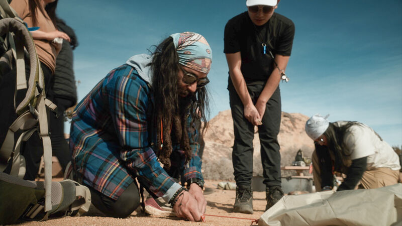 The image shows a group of people outdoors, possibly camping or hiking. One person is kneeling and appears to be working on something on the ground, perhaps setting up a tent. Another person is standing nearby, observing. There's a backpack visible, suggesting an outdoor activity. The setting seems to be a desert or arid environment with a clear sky.