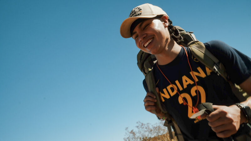 A young person with a backpack and a baseball cap smiles against a clear blue sky. They are wearing a dark blue t-shirt with "INDIANA 22" printed in gold. The person appears to be outdoors, possibly hiking, as suggested by the backpack and the sunny environment. The overall impression is one of joy and adventure.