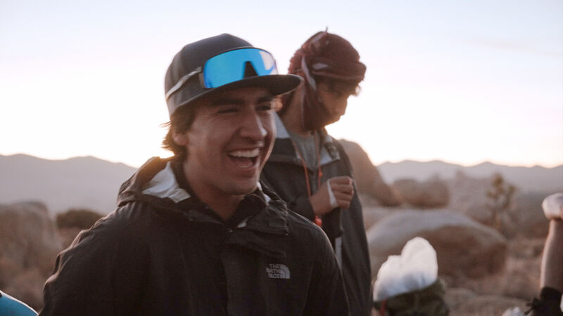The image shows a smiling man wearing a black jacket and a cap with blue-tinted sunglasses perched on top. He appears to be outdoors, possibly in a mountainous or desert region, given the background landscape. Another person is partially visible behind him, and there are other individuals or objects at the bottom of the frame, suggesting a group setting or activity.